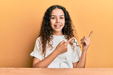 Gündelik kıyafetler giyen genç İspanyol kız masaya oturmuş gülümsüyor ve kameraya bakıyor. İki eliyle ve parmaklarıyla işaret ediyor.. 