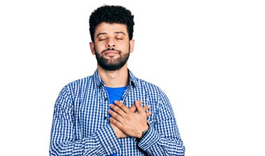 Sakallı, sıradan tişörtlü, göğsünde elleri göğsünde, yüzünde minnet dolu bir jest ile gülümseyen genç bir Arap. sağlık kavramı. 