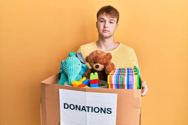 Kafkasyalı genç bir adam, elinde oyuncaklarla dolu bir bağış kutusu tutuyor. Yüzünde ciddi bir ifade var. Basit ve doğal bir şekilde kameraya bakıyorsun.. 