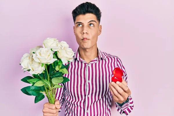 Young hispanic man holding bouquet of flowers and wedding ring making fish face with mouth and squinting eyes, crazy and comical.