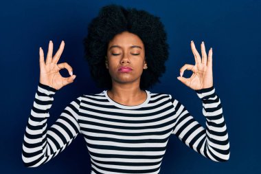 Genç Afro-Amerikalı kadın günlük kıyafetler giyip rahatlıyor ve gözleri kapalı gülümsüyor parmaklarıyla meditasyon hareketi yapıyor. yoga konsepti. 