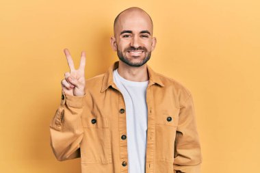 Gündelik kıyafetler giymiş genç kel bir adam. Gülümseyen mutlu bir yüzle kameraya bakıp zafer işareti yapıyor. İki numara.. 