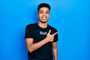 Genç, Afro-Amerikalı, personel tişörtü giymiş, neşeli, yüzünde bir gülümsemeyle, mutlu ve doğal bir ifadeyle elini ve parmağını kaldırmış bir adam. 