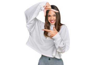 Balıkçı yaka kazak giyen genç esmer kadın gülümseyerek elleri ve parmakları gülen yüzlü bir çerçeve yapıyor. yaratıcılık ve fotoğrafçılık kavramı. 