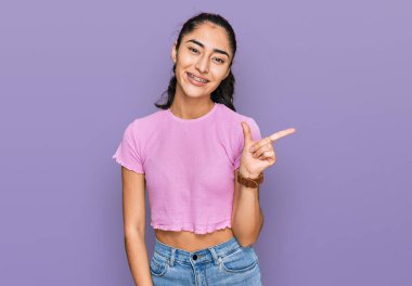 Diş teli takan, sıradan kıyafetler giyen, yüzünde kocaman bir gülümseme olan, eli kameraya bakan İspanyol bir genç kız.. 