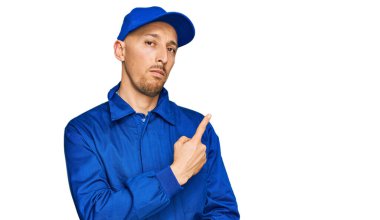 Sakallı, inşaatçı tulumu giymiş kel bir adam. El parmağı yanda, ciddi ve sakin bir yüz ifadesi var. 