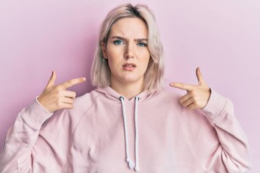 Genç sarışın kız parmaklarıyla ağzını işaret ediyor ve kafası karışık bir ifade takınıyor. şüphe kavramı. 