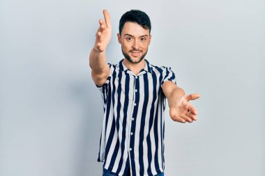 Gündelik giysiler giymiş genç İspanyol adam kameraya bakıp sarılmak için kollarını açmış gülümsüyor. Mutluluğu kucaklayan neşeli bir ifade.. 