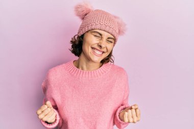 Şirin yün şapka takan genç esmer kadın çok mutlu ve heyecanlı, kolları havada, gülümseyen ve başarı için çığlık atan kazanan bir jest yapıyor. kutlama konsepti. 