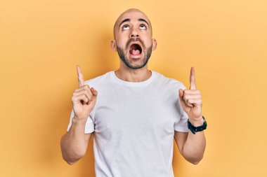 Gündelik beyaz tişört giyen genç kel adam şaşırmış ve şaşırmış bir şekilde yukarı baktı ve parmaklarıyla işaret etti ve kollarını kaldırdı.. 