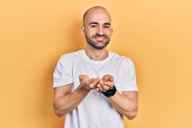 Elleri avuçlarında gülümseyen, günlük beyaz t-shirt giyen genç kel bir adam el hareketi yapıyor. Bekleme ve koruma 