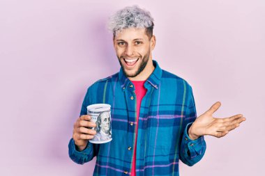 Modern boyalı saçları olan genç İspanyol bir adam kumbarayla başarıyı kutluyor mutlu bir gülümsemeyle ve elini kaldırarak kazanan bir ifadeyle. 