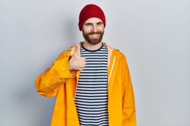 Sakallı, sarı yağmurluk giyen, mutlu bir el hareketi yapan beyaz bir adam. Kameraya bakıp başarıyı gösteren ifade. 