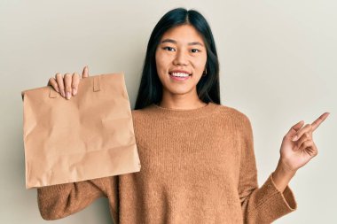 Elinde kağıt torba tutan genç Çinli kadın gülümsüyor mutlu bir şekilde elini ve parmağını yana doğru gösteriyor. 