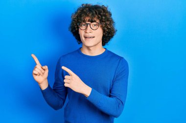 Yakışıklı genç adam günlük kıyafetler ve gözlük takıyor gülümsüyor ve kameraya bakıyor iki eliyle ve parmaklarıyla kenarı işaret ediyor.. 