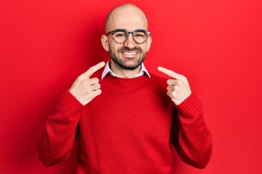 Gündelik kıyafetler ve gözlük takan genç kel adam neşeli bir şekilde gülümsüyor ve parmaklarıyla ve ağzıyla işaret ediyor. diş sağlığı kavramı. 