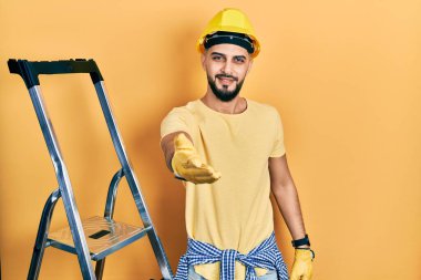 İnşaat merdivenlerinde sakallı yakışıklı bir adam. Kasketli, güler yüzlü, dostça bir selamlama ve hoş geldin hediyesi. başarılı iş. 