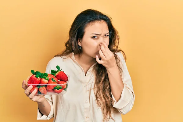 Young hispanic girl holding strawberries smelling something stinky and disgusting, intolerable smell, holding breath with fingers on nose. bad smell