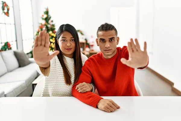 一对年轻的拉丁夫妇坐在圣诞树旁的桌子上 手心按着手掌 停止歌唱 用消极而严肃的手势警告人 — 图库照片