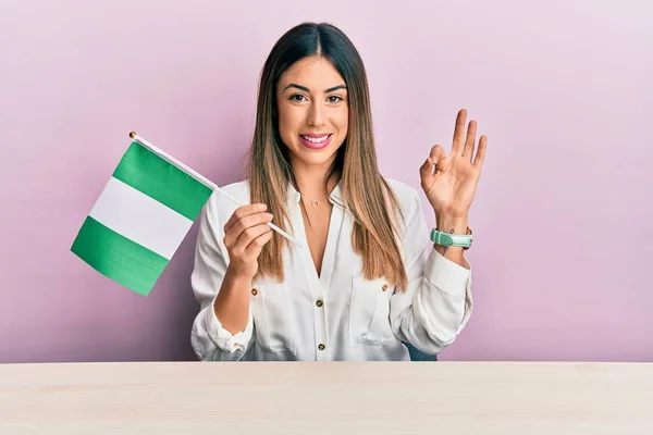 Jovem Hispânica Segurando Bandeira Nigéria Sentado Mesa Fazendo Sinal Com — Fotografia de Stock