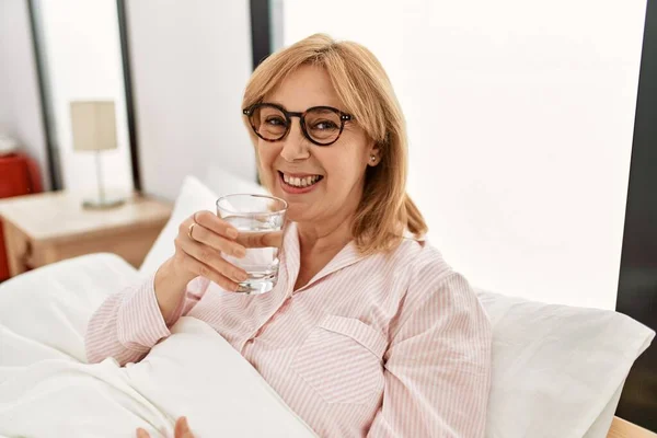 Blondynka Średnim Wieku Uśmiechnięta Szczęśliwa Szklanka Wody Leżąca Łóżku Domu — Zdjęcie stockowe