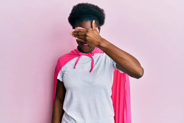 Young african american girl wearing superhero mask and cape costume smiling and laughing with hand on face covering eyes for surprise. blind concept.