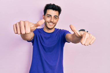 Gündelik tişört giyen genç İspanyol adam elinle olumlu jest yapmayı onaylıyor, gülümsüyor ve başarı için mutlu. kazanan jest. 