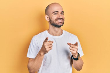 Sıradan beyaz tişört giyen genç kel adam mutlu ve komik yüzlü parmaklarını kameraya doğrultuyor. iyi enerji ve titreşimler. 