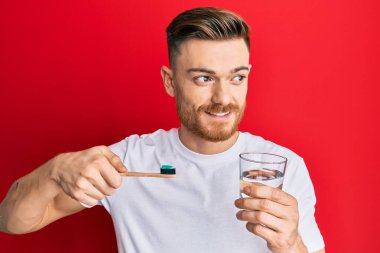 Diş fırçası ve bir bardak suyla elinde diş fırçası olan genç bir kızıl adam gülümsüyor ve başka yöne bakıyor.. 