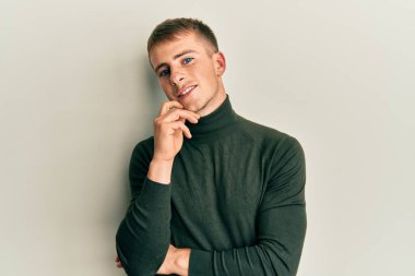 Gündelik giysiler giymiş genç beyaz bir adam kameraya kollarını kavuşturmuş ve çenesine dokunmuş bir şekilde gülümsüyor. Olumlu düşünmek. 