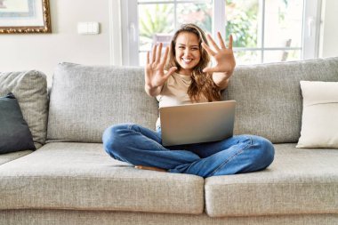 Güzel, genç, esmer bir kadın evdeki bilgisayar laptopunu kullanarak koltukta oturuyor. Kendini beğenmiş ve mutlu bir şekilde gülümserken on numarayı işaret ediyor.. 