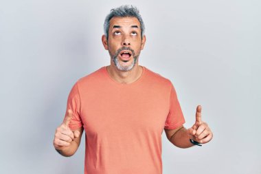 Gri tişörtlü yakışıklı orta yaşlı adam şaşkın şaşkın yukarı bakıyor ve parmaklarıyla işaret ediyor ve kollarını kaldırıyor.. 