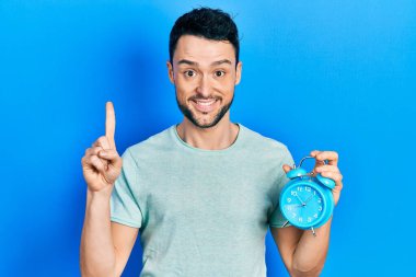 Çalar saati tutan genç İspanyol bir adam bir fikir ya da soruyla gülümsüyor ve mutlu bir yüzle işaret ediyor. 