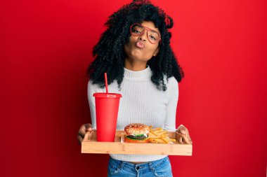 Afro saçlı Afro-Amerikalı bir kadın lezzetli bir klasik burger yiyor. Patates kızartması ve soda ile kameraya bakıp öpüşüyor. Güzel ve seksi. aşk ifadesi. 