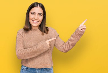 Genç, güzel bir kadın, günlük kıyafetler giyip gülümsüyor ve kameraya bakıyor. İki eliyle ve parmaklarıyla kenarı işaret ediyor.. 