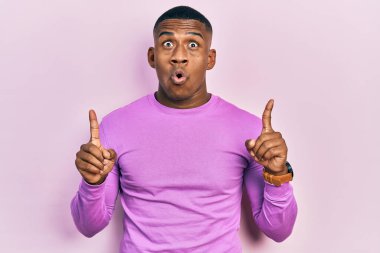 Gündelik pembe kazak giyen genç siyah adam şaşırmış ve şaşırmış bir şekilde yukarı bakıyor ve parmaklarıyla işaret ediyor ve kollarını kaldırıyor.. 