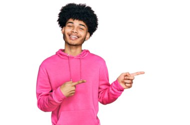 Afro saçlı, sıradan pembe kazak giyen, gülümseyen ve kameraya bakan, iki eli ve parmağı yanda olan Afro Amerikalı genç bir adam.. 