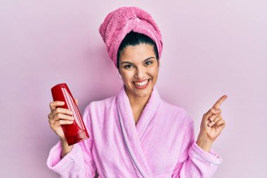 Duşlu bornoz giyen genç İspanyol kadın elinde şampuan, gülümsüyor. 