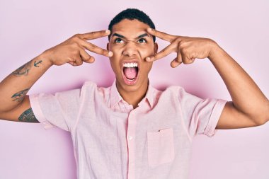 Genç Afrikalı Amerikalı adam zafer işareti ve barış hareketi yapıyor kızgın ve kızgın çığlıklar atıyor öfkeyle ve öfkeyle bağırıyor öfkeyle yukarı bakıyor. 