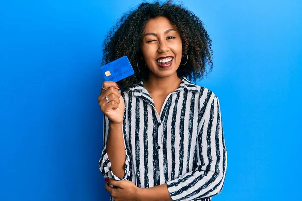 Beautiful african american woman with afro hair holding credit card winking looking at the camera with sexy expression, cheerful and happy face.
