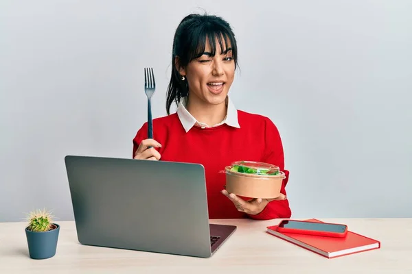 Young brunette woman with bangs working at the office eating healthy salad winking looking at the camera with sexy expression, cheerful and happy face.
