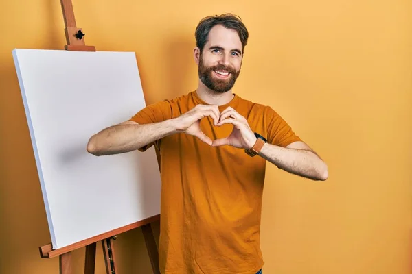 白种人留着胡子 站在画家的画架旁边 微笑着 用双手做着心形符号 浪漫的概念 — 图库照片