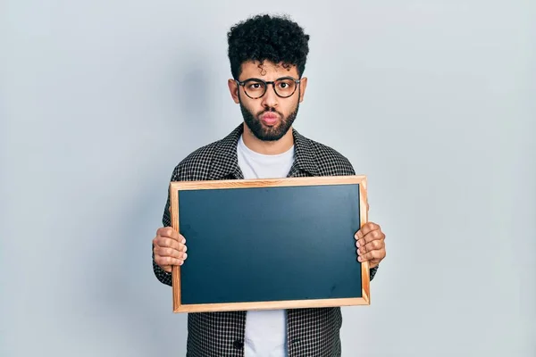 Young arab man with beard holding blackboard making fish face with mouth and squinting eyes, crazy and comical.