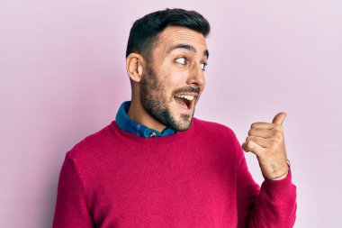 Gündelik kıyafetler giyen, gülümseyen, gülümseyen ve başparmağıyla yan tarafı gösteren İspanyol genç adam.. 