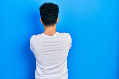 Sakallı, beyaz tişörtlü genç bir Arap. Arkaya bakıyor, kollarını kavuşturuyor. 