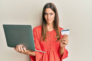 Genç ve güzel bir kadın dizüstü bilgisayar ve kredi kartı tutarken şüpheci ve gergin, sorun yüzünden kaşlarını çatıyor. negatif kişi. 