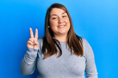 Güzel esmer ve cüsseli bir kadın. Günlük kıyafetler giyiyor. İki numarayı gösteriyor ve gülümsüyor.. 