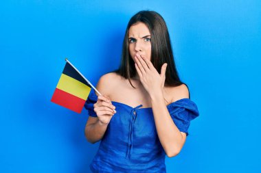 Belçika bayrağını elinde tutan genç esmer kız ağızlarını eliyle kapatıyor, şaşırmış ve hata yapmaktan korkuyor. şaşırmış ifade 