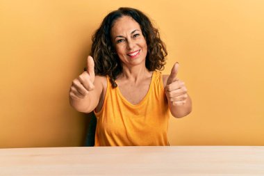 Güzel orta yaşlı kadın, gündelik giysiler içinde masada oturmuş, olumlu jestler yapıyor, gülümsüyor ve başarı için seviniyor. kazanan jest. 