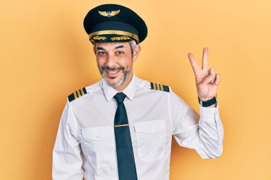 Yakışıklı, orta yaşlı, gri saçlı, uçak pilotu üniforması giyen, kameraya gülümseyen ve parmaklarıyla zafer işareti yapan bir adam. İki numara.. 